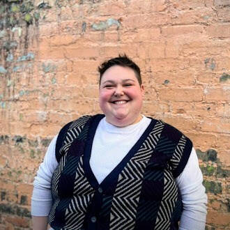 Olivia, with short dark hair and in a black striped vest over a long-sleeve white shirt, stands in front of a light brown brick wall